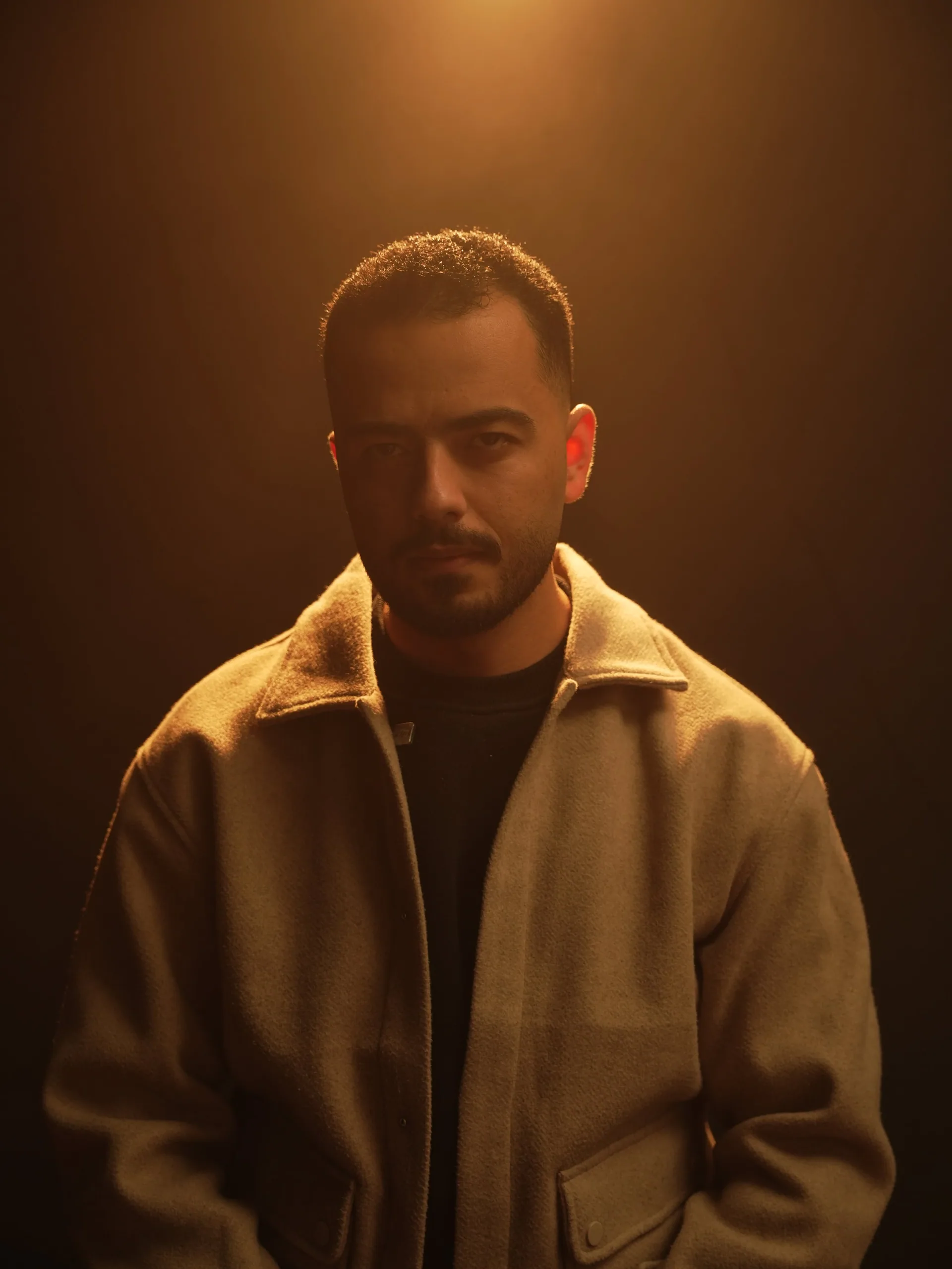 Portrait of Omar Diab in a beige jacket, lit by a warm amber light against a dark studio background